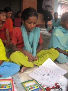 Girls sit on classroom mats with their school materials, listening to the lesson.